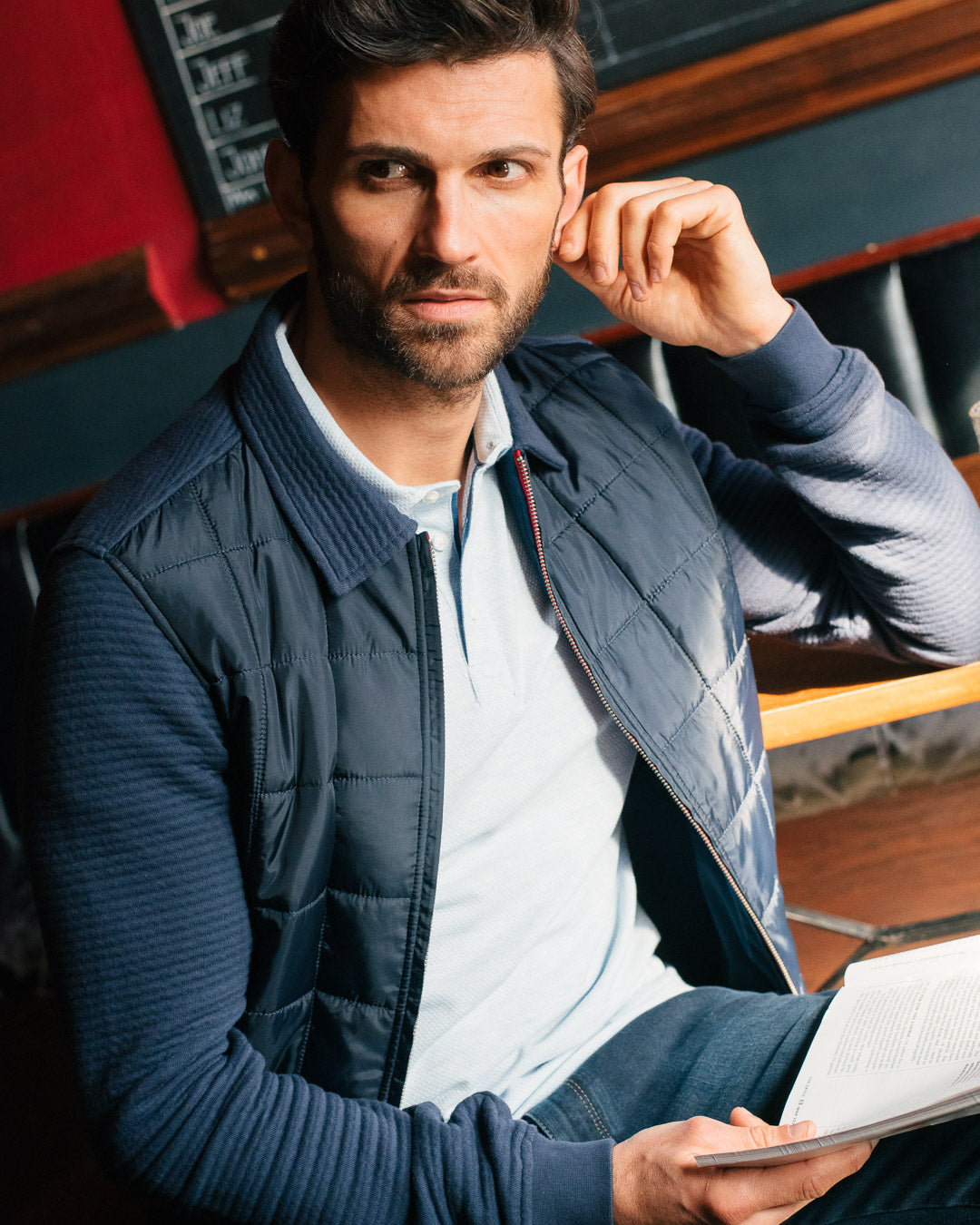  Image of a man in a casual pose, wearing a blue sweatshirt and a light blue polo shirt. He appears relaxed and comfortable, with a slight smile on his face, and is holding a notebook. Get inspired by Guide London's premium clothing collection for men's fashion.