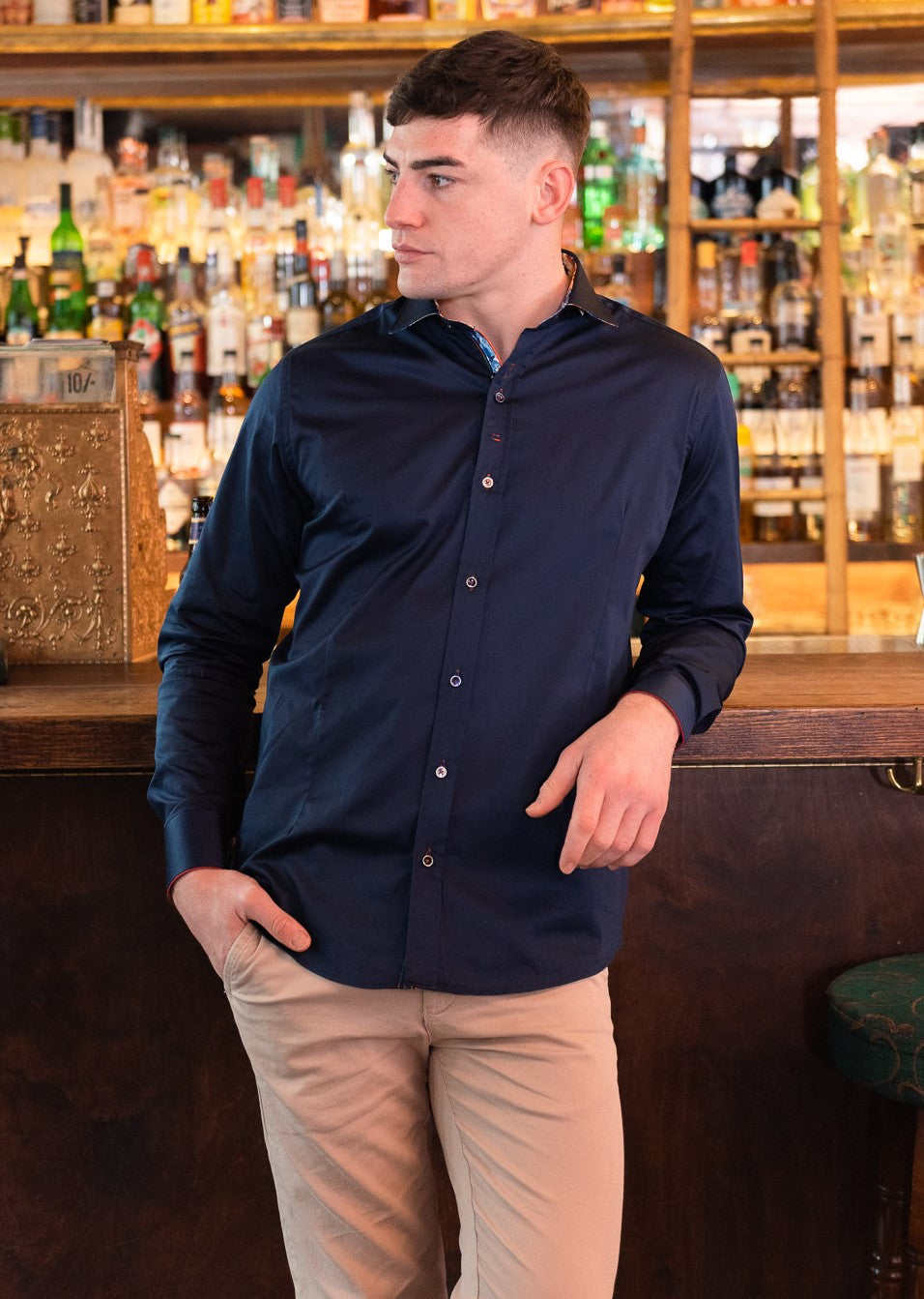 Image of a man wearing a navy blue long sleeve shirt and beige pants, standing in a casual pose at a bar. He appears relaxed and comfortable, with a slight smile on his face. Get inspired by Guide London's premium clothing collection for men's fashion.